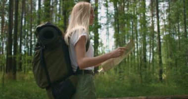 Elinde harita olan genç bir kadın ormanda ağır çekimde sırt çantasıyla dolaşıyor. Ormanda şortlu bir gezgin.