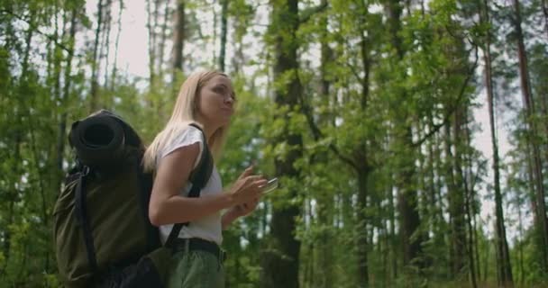 Une jeune femme avec un téléphone portable se promène dans la forêt en voyageant avec un sac à dos au ralenti. Voyageur en short dans les bois à la recherche de satellites GPS — Video