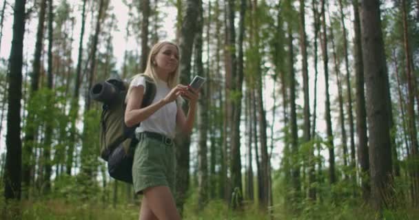 Молодая женщина с мобильным телефоном ходит по лесу с рюкзаком в замедленной съемке. Перемещение по лесу с помощью навигатора в мобильном телефоне — стоковое видео