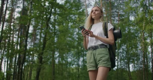 Een jonge vrouw met een mobiele telefoon loopt in slow motion door het bos met een rugzak. Reiziger in korte broek in het bos op zoek naar GPS satellieten — Stockvideo