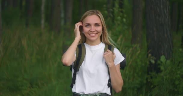 Retrato de una joven viajera bajo el sol mirando directamente a la cámara y coqueteando sonriente, Viajero con una mochila en el parque y en el bosque en cámara lenta — Vídeos de Stock