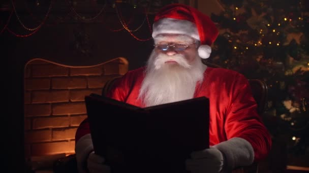 Homme âgé barbu en costume clause Père Noël feuilletant des pages de livre avec couverture rouge dans une pièce décorée thématiquement concept esprit de Noël close up. — Video