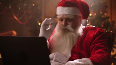 Modern Noel Baba. Neşeli Noel Baba dizüstü bilgisayarda çalışıyor ve gülümsüyor. Arka planda şöminesi ve Noel Ağacı olan sandalyesinde oturuyor..