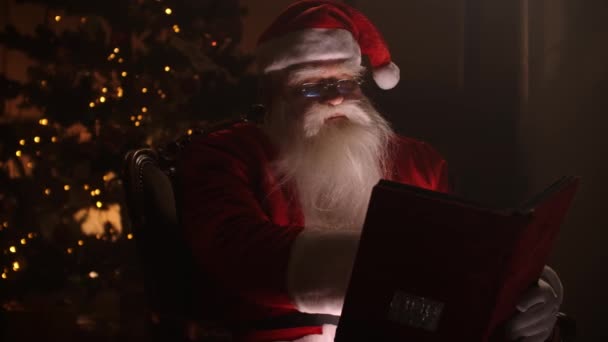 Radosny Mikołaj klauzula siedzi w swoim rocker w urządzonym pokoju, czytając książkę z czerwoną okładką - świąteczny nastrój, koncepcja ducha Bożego Narodzenia. — Wideo stockowe