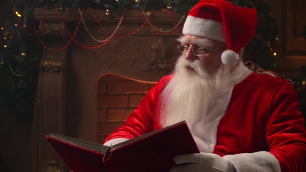 Alegre cláusula de santa sentado en su rockero en la habitación decorada, la lectura de un libro con cubierta roja - el estado de ánimo de vacaciones, el espíritu de Navidad concepto. — Vídeos de Stock