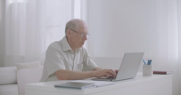 Anciano especialista masculino está trabajando desde casa, escribiendo y enviando correo electrónico por Internet, utilizando el ordenador portátil — Vídeo de stock