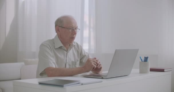 Ηλικιωμένος άνδρας δικηγόρος συμβουλεύεται τον πελάτη μέσω διαδικτύου, χρησιμοποιώντας φορητό υπολογιστή με web camera, κάθεται μόνος στο γραφείο του — Αρχείο Βίντεο
