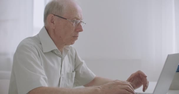 メガネをかけた高齢の灰色の男はノートパソコンを操作しネットサーフィンをしたりオンラインでメッセージでチャットしたりキーボードを押したり — ストック動画