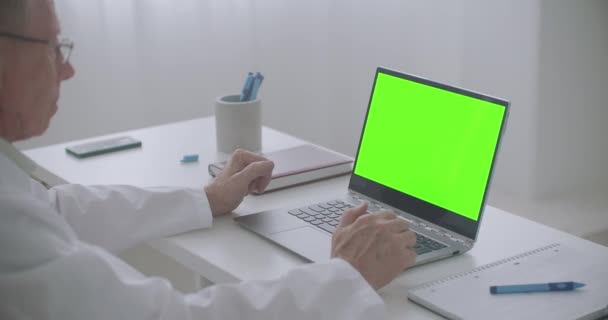 Médico masculino está consultando en línea, mirando la pantalla verde de la computadora portátil en la oficina de la clínica, tecnología de croma key — Vídeos de Stock