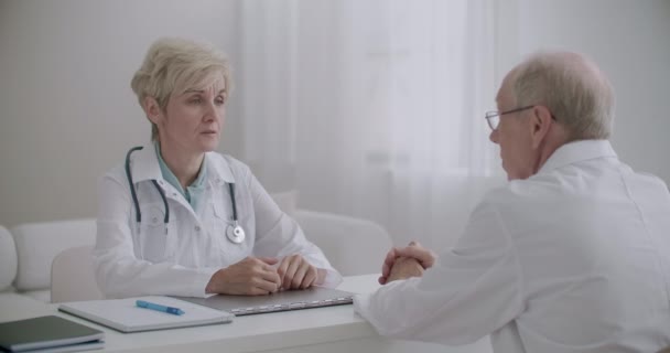 Ηλικιωμένος άνδρας θεραπευτής συμβουλεύεται γυναίκα επικεφαλής ιατρός του νοσοκομείου στο γραφείο, συζητά και αποφασίζει — Αρχείο Βίντεο