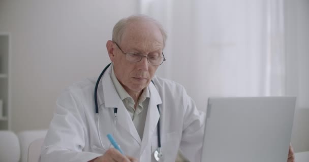 Professor idoso de medicina está visualizando webinar por laptop no escritório da clínica e fazendo anotações, e-learning para profissionais de saúde — Vídeo de Stock