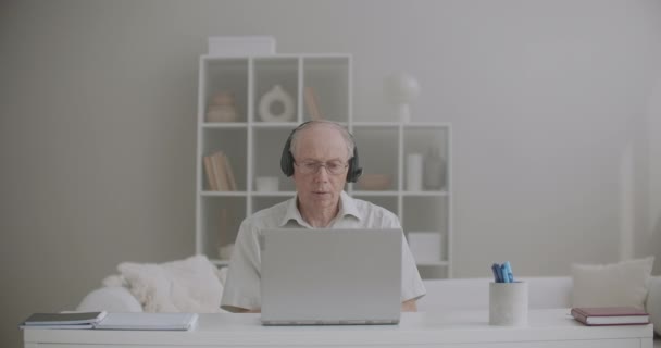 高齢の男性教師は講義をオンラインで行い家にいますインターネット接続とヘッドフォンでノートパソコンを使います — ストック動画