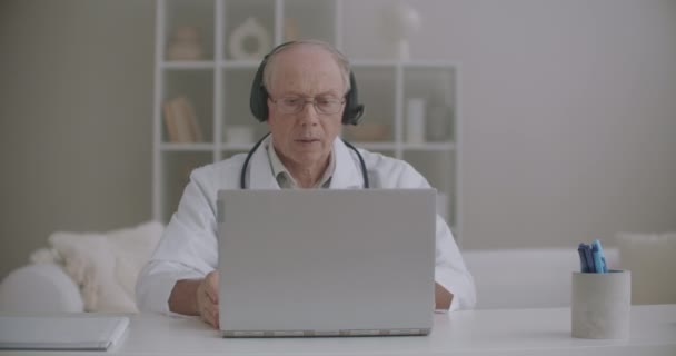 男性医師はオンラインの同僚や患者に相談しています高齢男性セラピストはクリニックに座ってオンラインでチャットしています — ストック動画