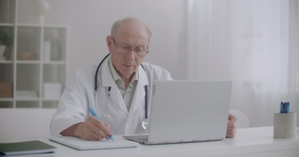 Médico idoso está ouvindo paciente na consulta on-line, olhando para a tela do laptop e escrevendo notas no papel — Vídeo de Stock