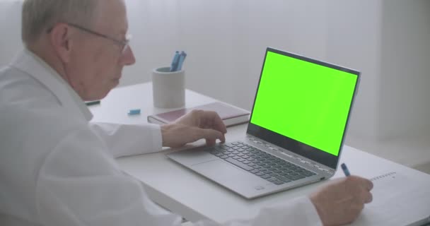 Elderly male doctor is reading article in internet, looking at chroma key display of laptop and writing — Stock Video