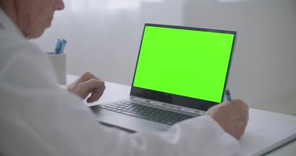 Medico sta cercando sul computer portatile con schermo a chiave cromatica sul tavolo in ufficio della clinica, scrivendo note — Video Stock