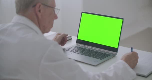 Laptop met groen scherm op tafel van werkende arts, man kijkt naar display en het schrijven van notities, e-learning concept — Stockvideo