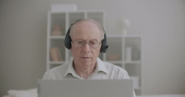 ヘッドフォンを装着した高齢者がノートパソコンのディスプレイを見て自宅からオンラインで講義をしています — ストック動画