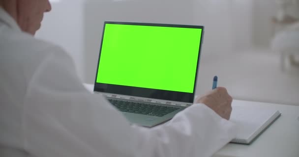Il medico sta cercando sul computer portatile con schermo verde sul tavolo in ufficio della clinica, scrivendo note — Video Stock