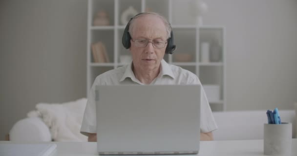 Profesor varón de edad se está comunicando por videollamada desde casa, utilizando el ordenador portátil y auriculares — Vídeo de stock