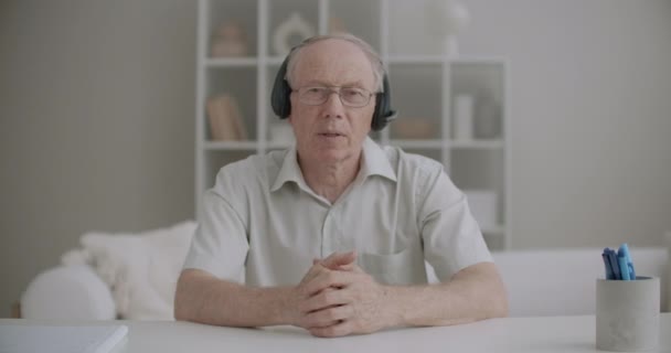 Homem idoso com fones de ouvido está olhando para câmera, cumprimentando o público on-line e começando a aprender webinar — Vídeo de Stock
