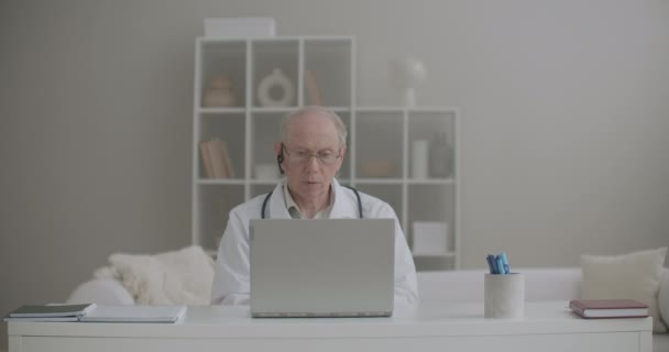 Ο ηλικιωμένος άνδρας ιατρός εργάζεται εξ αποστάσεως από το γραφείο του στην κλινική, χρησιμοποιώντας φορητό υπολογιστή για επικοινωνία με συναδέλφους — Αρχείο Βίντεο