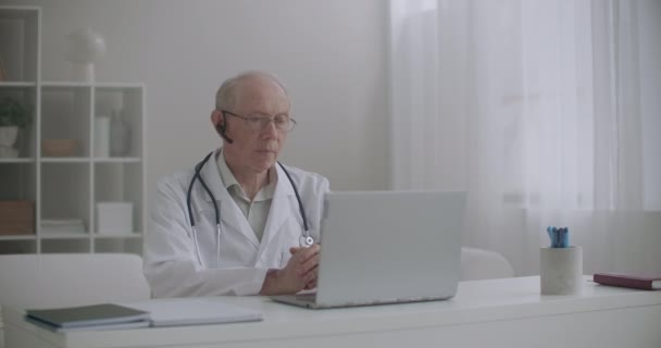 Oudere mannelijke arts raadpleegt patiënt online, zit op kantoor van de kliniek en communiceert via internet op laptop — Stockvideo