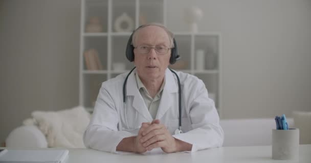 白人のガウンを着た高齢男性医師,眼鏡とヘッドフォンはオンラインで通信しています,前頭葉の肖像画 — ストック動画