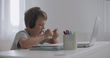 Küçük çocuk internette mikrofonlu kulaklıkla sohbet ediyor, dizüstü bilgisayara ve çizime bakıyor, evde masada oturuyor.