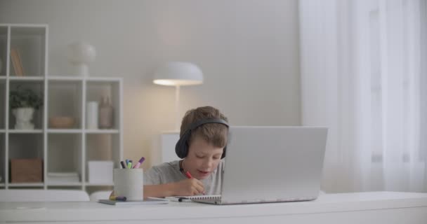 Niño de la escuela está escuchando cuento de hadas a través de auriculares por cuaderno y dibujo, sentado a la mesa solo en la habitación — Vídeo de stock