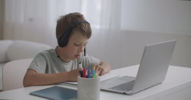 小男孩正在笔记本电脑上为孩子们观看教育视频，并通过耳机听写在纸上 — 图库视频影像