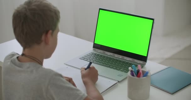 Bambino sta disegnando seduto a tavola con computer portatile con schermo verde per la tecnologia chroma key, bambino a casa nel fine settimana — Video Stock