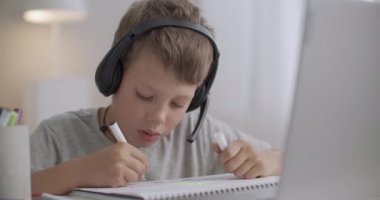 Komik okul çocuğu, uzaktan öğrenme oturumunda kulaklıkla öğretmenini dinliyor ve defter içinde resim çiziyor.