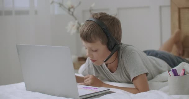 Niño pequeño está dibujando acostado en la cama en su habitación y la comunicación en línea por cámara web y auriculares — Vídeos de Stock