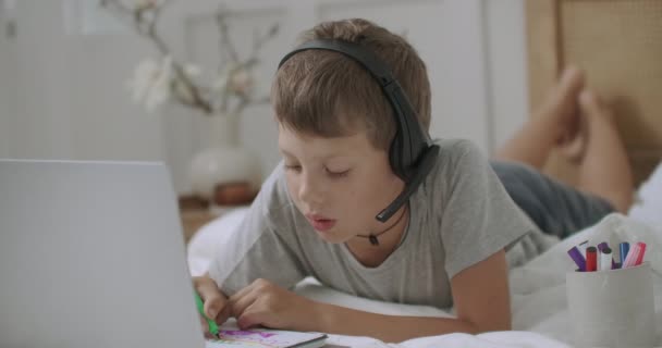 Bambino sta visualizzando cartoni animati o film per bambini da computer portatile e disegno, sdraiato sul letto nella sua stanza — Video Stock