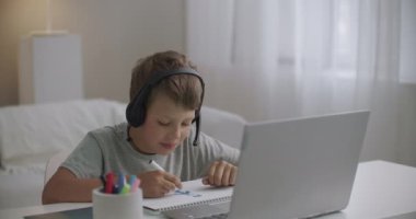 Öğrenci çocuk, öğrenme videosunu defterden izliyor ve kulaklıkla öğretmenini dinliyor.