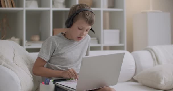 어린 소년은 소파에 앉아 종이에 그림을 그리고 있고, 헤드폰으로 비디오 테이프를 듣는 노트북을 사용하고 있습니다 — 비디오