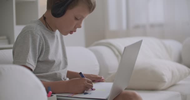 Bambino ragazzo sta disegnando e dipingendo immagini in album e chattare online da cuffie con microfono, stare a casa — Video Stock