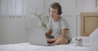 Neşeli çocuk bilgisayarda biriyle görüntülü iletişim kuruyor, kulaklık takıyor, evde yatakta oturuyor.