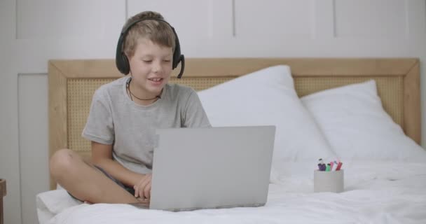 Menino está sentado na cama em casa e conversando online por notebook, falando no microfone de fones de ouvido — Vídeo de Stock