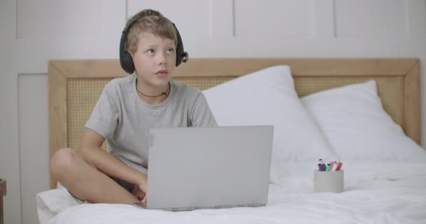 Niño pequeño está charlando con los padres en línea por ordenador portátil con auriculares del campamento de verano, sentado en la cama en la habitación — Vídeos de Stock