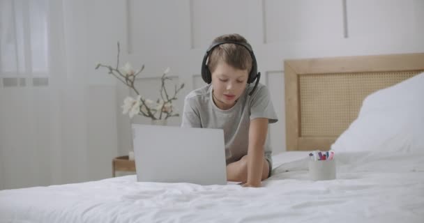 Internetsüchtiger kleiner Junge nutzt Laptop zum Kommunizieren und Spielen, sitzt im Bett und zeichnet auf Papier — Stockvideo