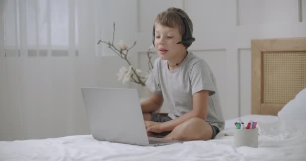 Niño alegre se está comunicando con alguien por videollamada en el ordenador portátil, el uso de auriculares, sentado en la cama en casa — Vídeo de stock