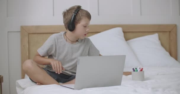 Przedszkolak rysuje i rozmawia online z przyjacielem lub rodzicem przez laptop ze słuchawkami, siedzi w domu — Wideo stockowe