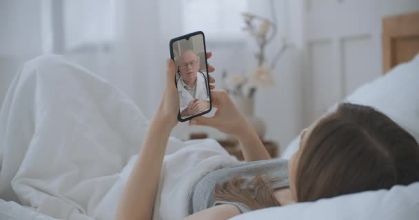 Female medical assistant wears white coat, headset video calling distant patient on smartphone. Doctor talking to client using virtual chat telephone app. Telemedicine, remote healthcare services — Stock Video