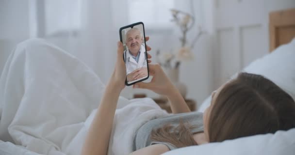 Jeune fille allongée au lit à la maison en utilisant Smartphone pour parler à son médecin via l'application médicale de vidéoconférence. Belle femme vérifie les symptômes possibles avec un médecin professionnel, en utilisant le chat vidéo en ligne. — Video