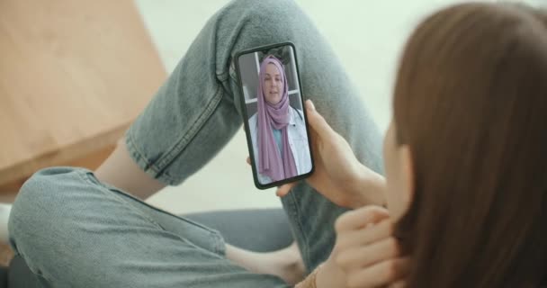 Mujer que utiliza la aplicación médica en la consulta de teléfonos inteligentes con la mujer árabe musulmana en el médico de hijab a través de videoconferencia. Mujer usando el chat en línea para hablar con la familia terapeuta y pandemia de coronavirus. — Vídeos de Stock
