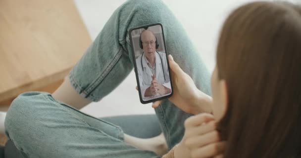 Asistente médica femenina usa bata blanca, auriculares video llamadas paciente distante en el teléfono inteligente. Doctor hablando con el cliente utilizando la aplicación de teléfono de chat virtual. Telemedicina, servicios de salud a distancia — Vídeos de Stock