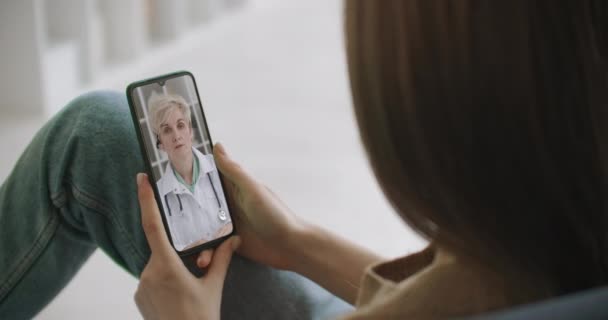 Kvinna kontrollerar eventuella symtom med professionell läkare, med hjälp av online videochatt. Ung flicka sjuk hemma med hjälp av smartphone för att prata med sin läkare via video konferens medicinsk app. — Stockvideo