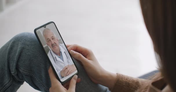 Kvinnelig medisinsk assistent bruker hvit frakk, hodetelefon video kaller pasient på smarttelefon. Legen snakker med klienten via en virtuell chat-app. Telemedisin, fjernhelsetjenester – stockvideo